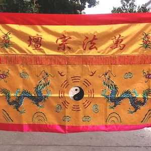 May include: A rectangular, gold-colored banner with red and pink borders. The banner features embroidered dragons, Chinese characters, and a yin-yang symbol. Decorative fringe is along the bottom of the banner.