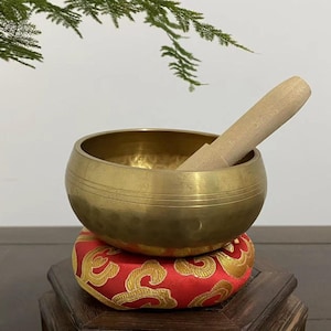 May include: A brass singing bowl with a wooden striker rests on a red cushion with gold embroidery. The bowl is round with horizontal lines and sits on a small wooden stand. A sprig of greenery is in the background.