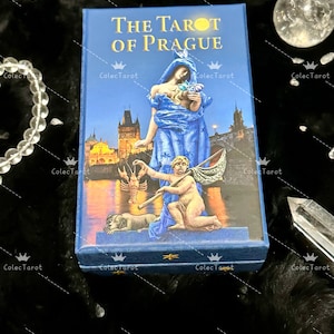 May include: The Tarot of Prague deck box features a blue background with a depiction of a woman in a blue dress standing on a bridge over a river. The woman is holding a flower and a small child is standing next to her. The box is decorated with gold stars and the text "The Tarot of Prague".