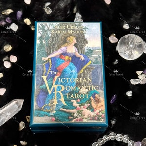 May include: The Victorian Romantic Tarot deck box features a blue background with gold lettering and a detailed illustration of a woman in a flowing gown sitting on a throne. The box is surrounded by crystals and a bracelet.