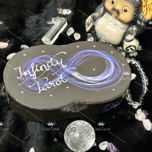 May include: A black box with a white infinity symbol and the words "Infinity Tarot" on the front. The box is sitting on a black fuzzy surface.