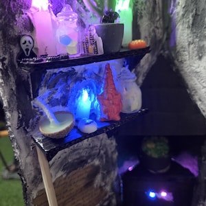 May include: A miniature Halloween diorama with shelves displaying candles, a cactus, a jar with colourful cubes, and a small Ghostface figure. The scene is lit with purple and blue lights, creating a spooky atmosphere. The diorama has a cave-like appearance.