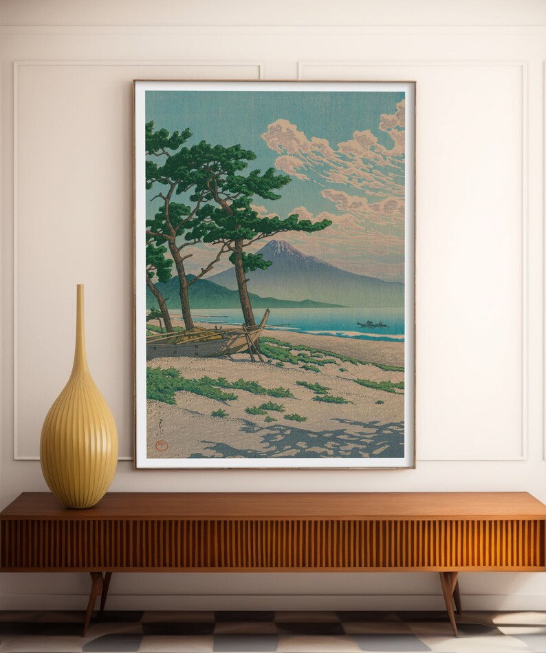 May include: A vintage Japanese woodblock print depicting a mountain range, a body of water, a boat on the shore, and two pine trees. The print is framed in a white frame.