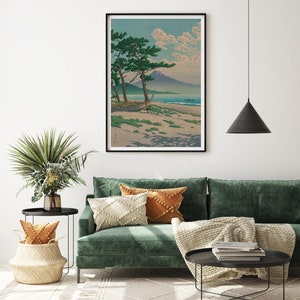 May include: A green velvet sofa with a white and orange throw blanket and pillows. A black coffee table with books and a black end table with a wicker basket and a potted plant are in the foreground. A framed print of a Japanese landscape with a mountain and a beach is hanging on the wall.