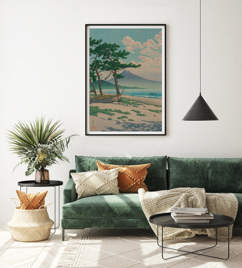 May include: A green velvet sofa with a white and orange throw blanket and pillows. A black coffee table with books and a black end table with a wicker basket and a potted plant are in the foreground. A framed print of a Japanese landscape with a mountain and a beach is hanging on the wall.
