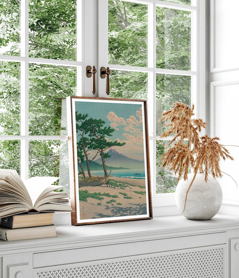 May include: A framed print of a Japanese landscape painting depicting a mountain, a boat on a beach, and two pine trees. The print is in a brown frame and is hanging on a white wall.