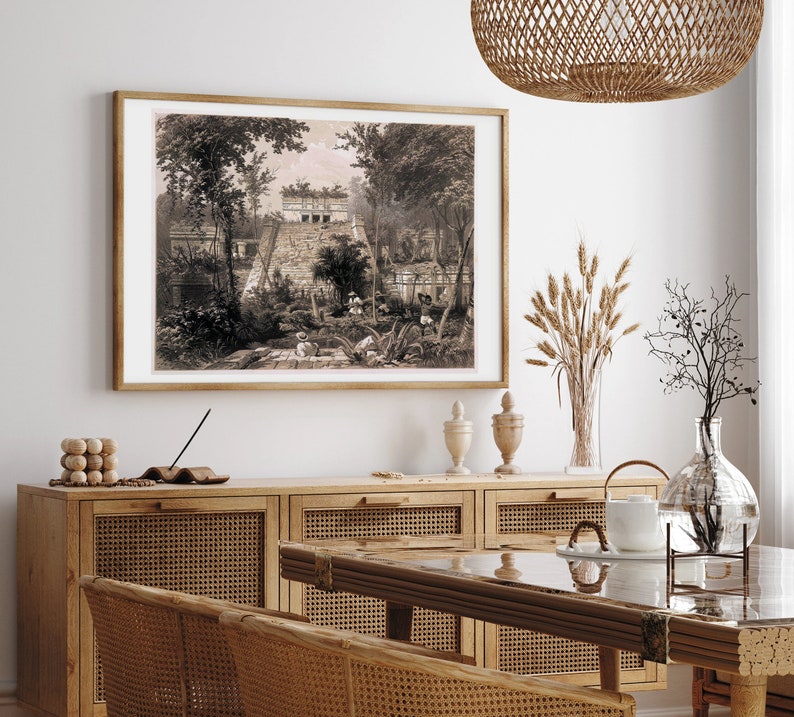 K&ouml;nnte beinhalten: Eine Vintage-Illustration einer Maya-Pyramide in einer Dschungelumgebung. Das Bild ist in einem braunen Holzrahmen gerahmt und h&auml;ngt an einer wei&szlig;en Wand &uuml;ber einem Holzschrank mit Rattan-T&uuml;ren.