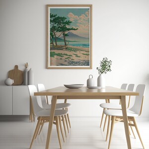 May include: A framed print of a Japanese landscape with a mountain in the distance, a beach in the foreground, and two pine trees in the middle ground. The print is in a light wood frame and is hanging on a white wall.