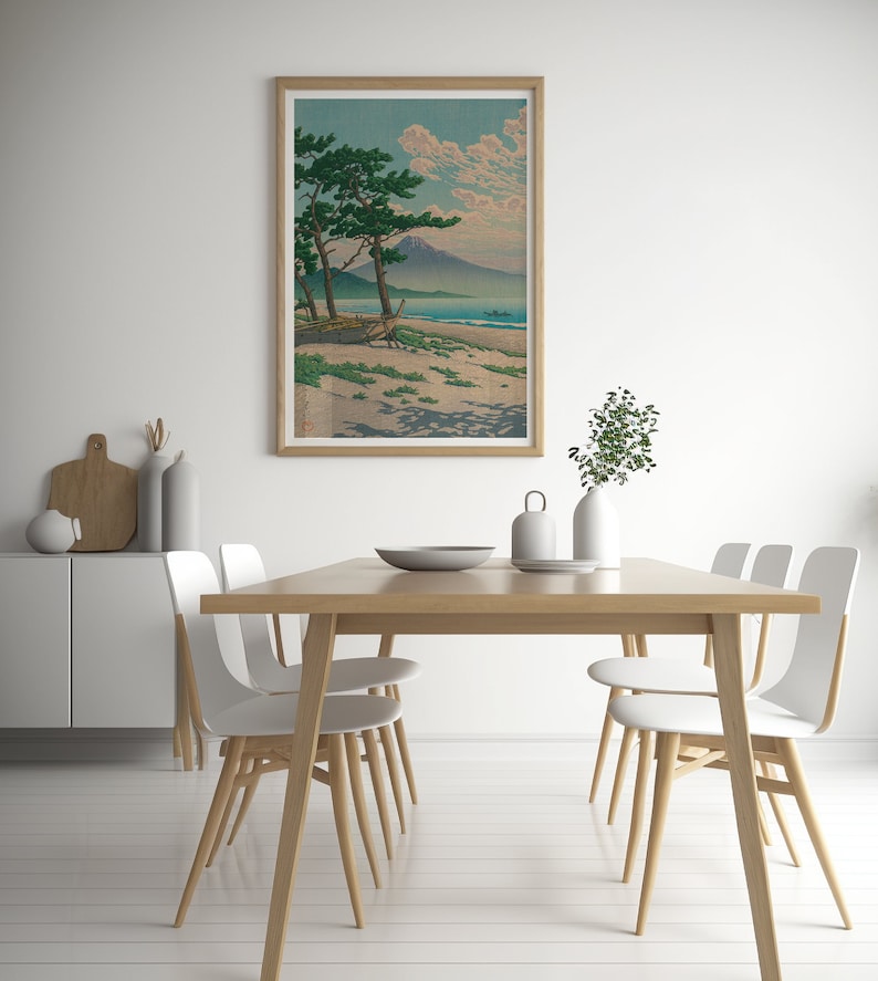May include: A framed print of a Japanese landscape with a mountain in the distance, a beach in the foreground, and two pine trees in the middle ground. The print is in a light wood frame and is hanging on a white wall.