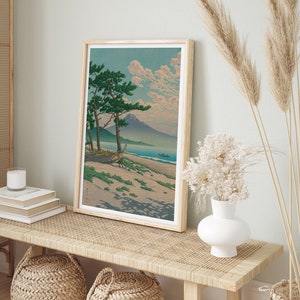 May include: A framed print of a Japanese landscape painting depicting a mountain, a beach, and two pine trees. The painting is in a light wood frame and is hanging on a wall.