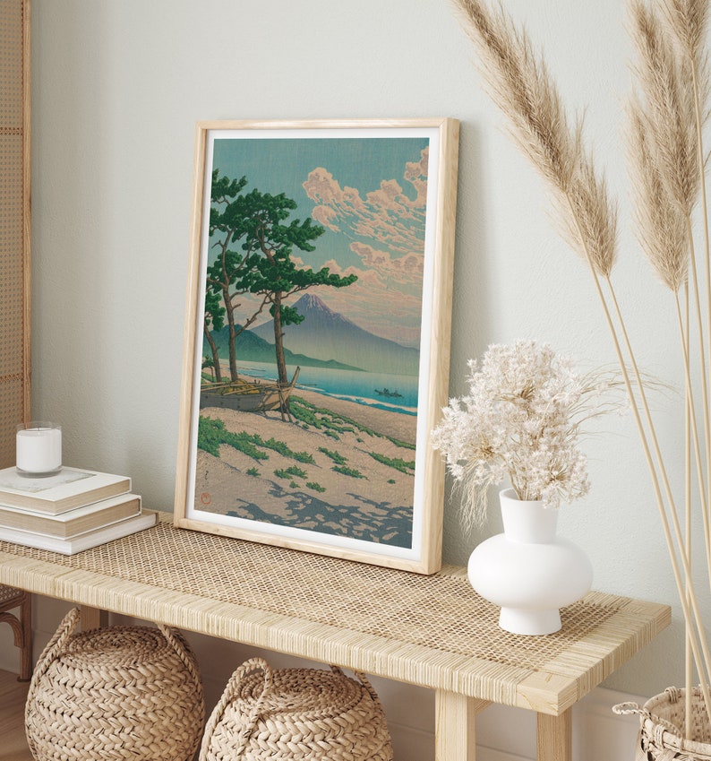 May include: A framed print of a Japanese landscape painting depicting a mountain, a beach, and two pine trees. The painting is in a light wood frame and is hanging on a wall.