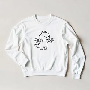 May include: White sweatshirt featuring a black and white cartoon dinosaur lifting weights. The dinosaur has a friendly expression. The design is centered on the front of the long-sleeved crew neck sweatshirt.