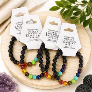 May include: Four stretch bracelets featuring black porous lava rock beads and colorful gemstone accents. Each bracelet is attached to a white display card labeled MYSTIC SHOWERS CHAKRA LAVA ROCK BRACELET with instructions to add 1-2 drops of oil.