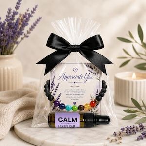 May include: A clear plastic gift bag tied with a black satin ribbon containing a chakra bracelet with black lava beads and colorful stones, and a small amber glass bottle labeled CALM. A card inside reads Appreciate You with lavender art.