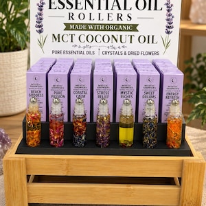 Può includere: Un espositore con sette flaconi in vetro con applicatore a sfera per oli essenziali, contenenti fiori secchi o oli di colori diversi. I flaconi sono disposti davanti a scatole viola etichettate Mystic Showers con un cartello sopra.