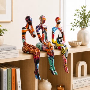 Pode incluir: Três esculturas abstratas em forma humana sentadas numa prateleira, cada uma com um padrão de graffiti multicolorido vibrante com salpicos arrojados de tinta vermelha, azul, amarela e preta cobrindo as suas formas suaves e estilizadas.