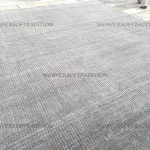 Peut inclure: Un grand tapis rectangulaire gris avec une surface texturée et côtelée. Le tapis présente un motif subtil et les mots "WEAVERSOFTRADITION" sont imprimés sur la surface. Le tapis semble être fait d'une matière douce.