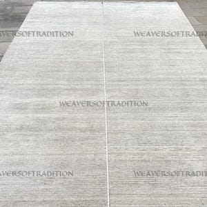 Peut inclure: Un grand tapis rectangulaire gris clair avec un motif à rayures subtiles. Le tapis est fait d'une matière douce et porte l'inscription "WEAVERSOFTRADITION". Le tapis est probablement utilisé pour la décoration intérieure.