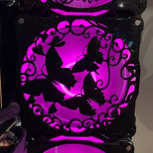 May include: A black computer fan with a decorative cutout of butterflies and floral designs. The fan is illuminated with a bright purple light.