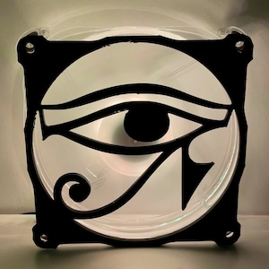 May include: A black and white computer fan with a stylised eye of Horus design. The fan is made of clear plastic and has a black frame.