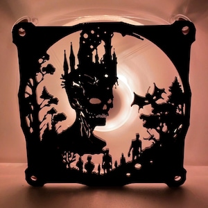 May include: Black metal fan with a silhouette of a creature with a crown, trees, and figures. The fan is illuminated from behind, creating a dramatic effect.
