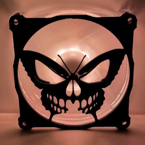 Puede incluir: Una cubierta de ventilador de computadora de plástico negro con un diseño de calavera y mariposa. La cubierta del ventilador está iluminada desde atrás, creando un efecto brillante.