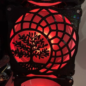May include: A black computer fan with a red light shining through a cut-out design of a tree with roots and a geometric pattern.