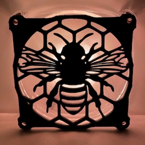 May include: Black metal cutout of a bee with wings spread, inside a honeycomb design. The bee is silhouetted against a light background.