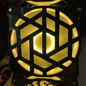 May include: A black computer fan with a geometric design and a yellow light shining through the centre.