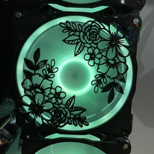 Puede incluir: Un ventilador de ordenador negro con una luz verde y un diseño floral. El diseño está recortado del ventilador y es negro.