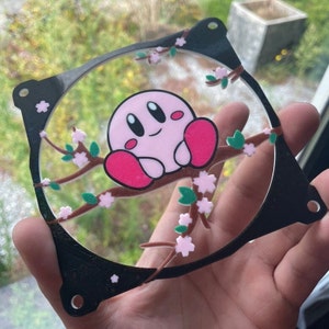 Könnte beinhalten: Ein schwarzer Plastik-Lüftergrill mit einer rosa und weißen Comicfigur, die auf einem Ast mit rosa Blüten sitzt. Die Figur hat einen rosa Körper, rote Arme und ein weißes Gesicht mit schwarzen Augen.