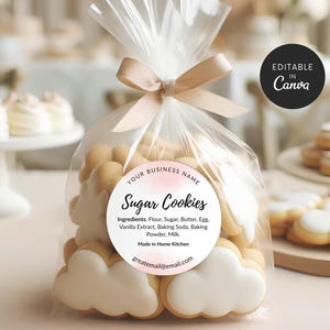 May include: A transparent cellophane bag tied with a beige ribbon, filled with sugar cookies. The cookies are cloud-shaped and round, with white icing. A circular label reads "Sugar Cookies" and lists ingredients.