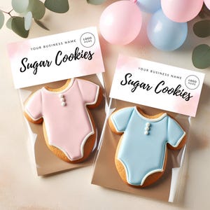 May include: Two baby onesie-shaped sugar cookies, one pink and one blue, in clear cellophane bags. Each cookie has white icing details and three small white buttons. The bags are topped with a label that reads "Sugar Cookies" and space for a business name and logo.