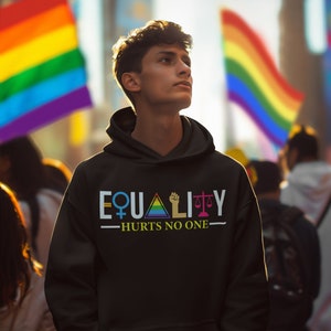 Könnte beinhalten: Eine Person trägt einen schwarzen Hoodie mit dem Text "EQUALITY - HURTS NO ONE" und Symbolen, die für Gleichstellung der Geschlechter stehen, ein Regenbogen-Dreieck, eine geballte Faust und eine Waage.