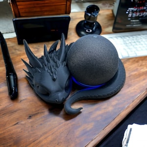 Könnte beinhalten: Eine schwarze Drachenfigur mit einem blauen Licht, die auf einem schwarzen intelligenten Lautsprecher ruht. Der Lautsprecher steht auf einem Holztisch.