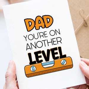 May include: A white greeting card with the words "DAD YOU'RE ON ANOTHER LEVEL" in orange and black. The card features an illustration of a level tool. The card is held by two hands.