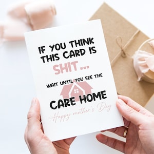 Two hands holding a cheeky and funny Mother's Day card with a sarcastic care home joke over a minimalist white desk.