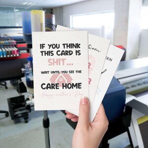 A hand holding up a funny care home joke Mother's Day card alongside other custom greeting cards in a printing workshop with a professional printer in the background.
