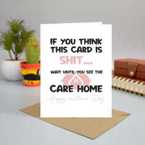 A funny and sarcastic Mother's Day card featuring a care home joke, standing upright on a desk next to a brown envelope, a small potted cactus, and a spiral notebook.