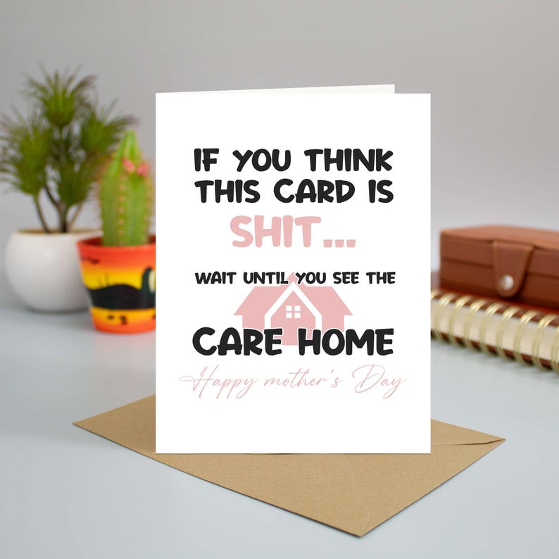 A funny and sarcastic Mother's Day card featuring a care home joke, standing upright on a desk next to a brown envelope, a small potted cactus, and a spiral notebook.