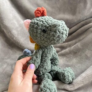 May include: A handmade, plush crochet dinosaur in shades of green, with a colorful crest of orange, pink, yellow, and blue. The dinosaur has a textured, soft appearance and a black button eye. The toy is sitting on a gray blanket.