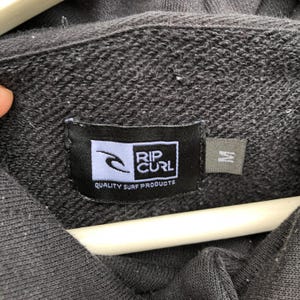 May include: Close-up of a dark gray hoodie with the "RIP CURL" logo label. The label is black with a white logo and text that reads "QUALITY SURF PRODUCTS". A size "M" tag is visible.