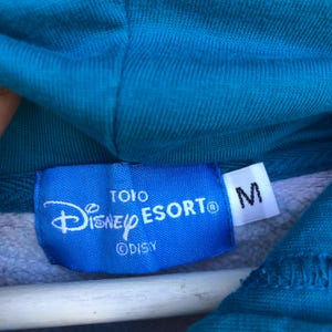 May include: Close-up of a blue hoodie with a Disney Esort label. The label is blue with white text and the Disney logo. A white tag with the letter "M" is attached.