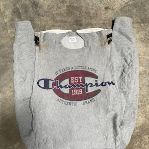 May include: A gray Champion sweatshirt with the text "IT TAKES A LITTLE MORE" above the Champion logo. The logo is in a dark blue and red color scheme, with "EST 1919" in the center. The words "AUTHENTIC BRAND" are below the logo.