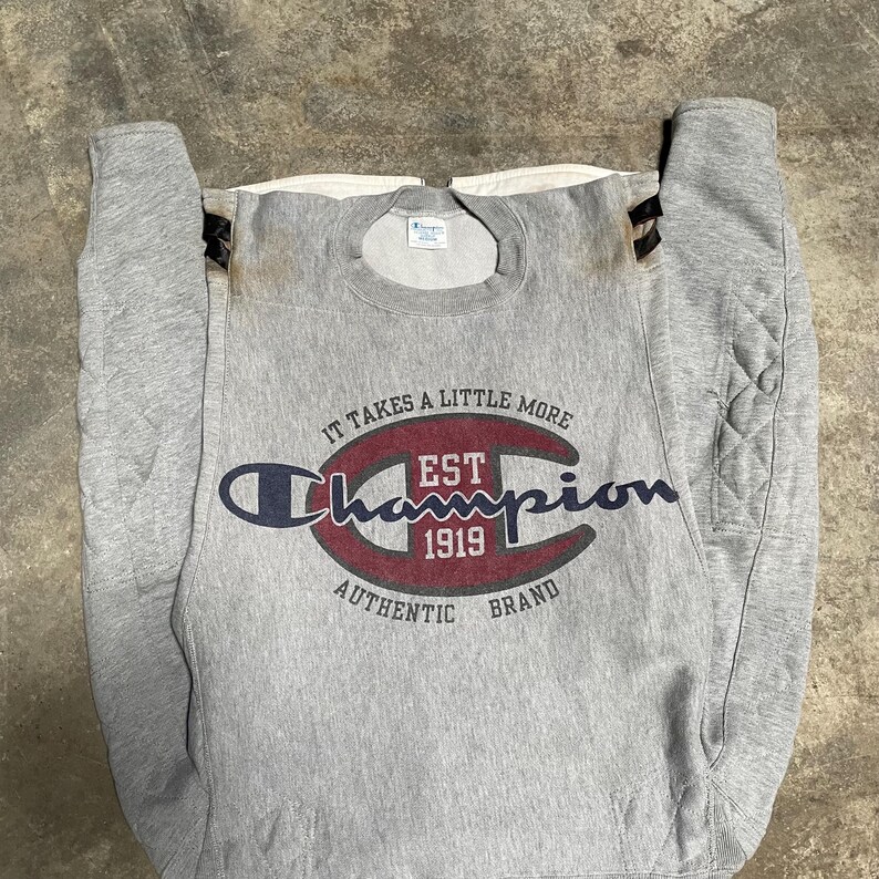 May include: A gray Champion sweatshirt with the text "IT TAKES A LITTLE MORE" above the Champion logo. The logo is in a dark blue and red color scheme, with "EST 1919" in the center. The words "AUTHENTIC BRAND" are below the logo.