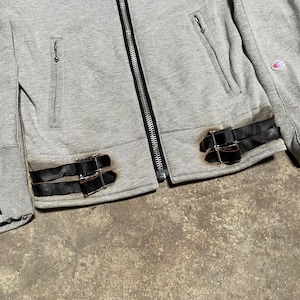 May include: A gray zip-up jacket with black leather straps and silver buckles. The jacket has two front pockets with zippers and a Champion logo on the sleeve. The jacket is made of a soft, comfortable material.