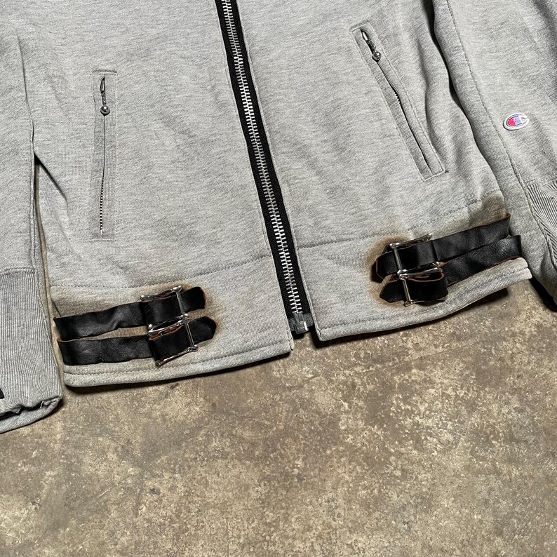 May include: A gray zip-up jacket with black leather straps and silver buckles. The jacket has two front pockets with zippers and a Champion logo on the sleeve. The jacket is made of a soft, comfortable material.