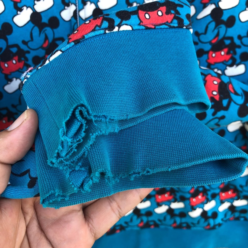 May include: A close-up of a blue sweatshirt sleeve with a Mickey Mouse pattern. The sleeve cuff is a darker blue and shows signs of wear. The background is filled with a repeating pattern of Mickey Mouse.