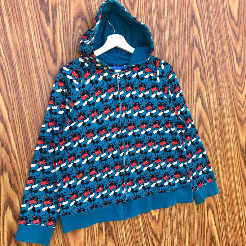 May include: Teal zip-up hoodie featuring a repeating pattern of Mickey Mouse. The hoodie has a hood, a full-length zipper, and teal cuffs and hem. The Mickey Mouse pattern includes red shorts, black ears, and white gloves.