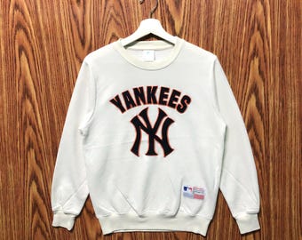 May include: White crew neck sweatshirt featuring the text "YANKEES" above a black and navy blue "NY" logo. The sweatshirt has long sleeves and a small tag on the lower left side. The garment is hanging on a white hanger.